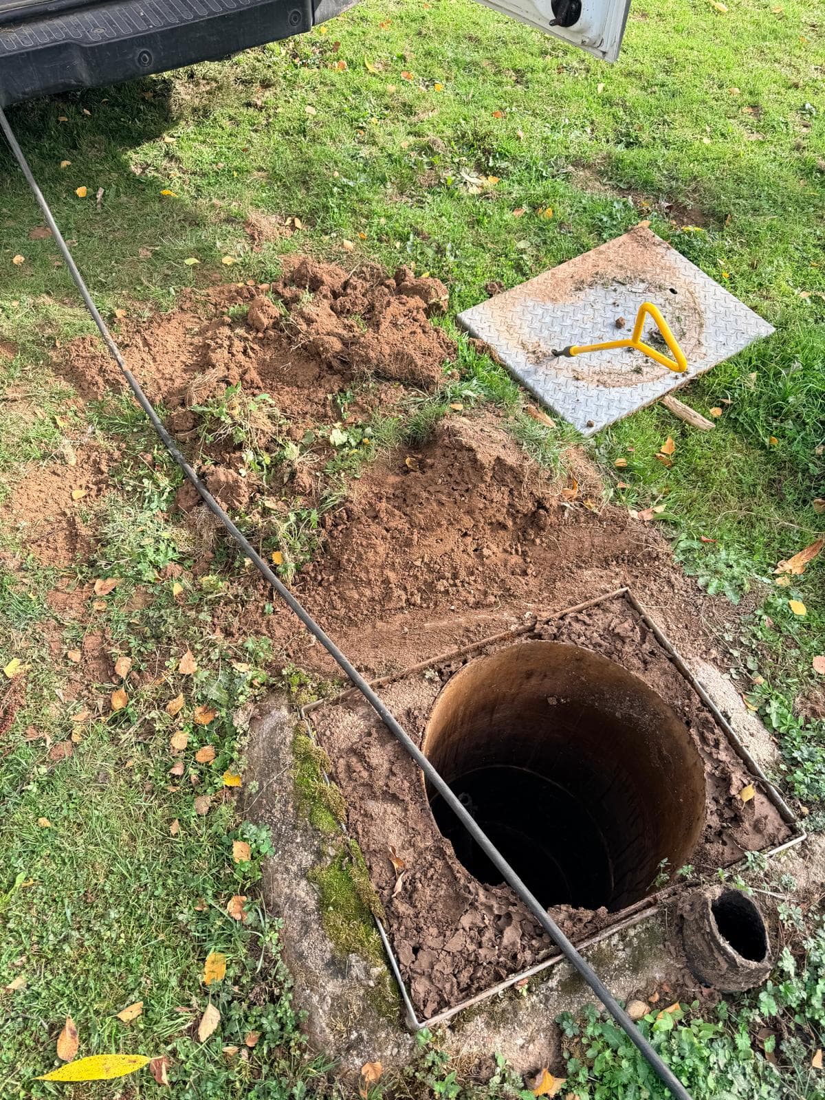 Uncovered circular septic-system access opening in grass, with the lifted cover alongside it